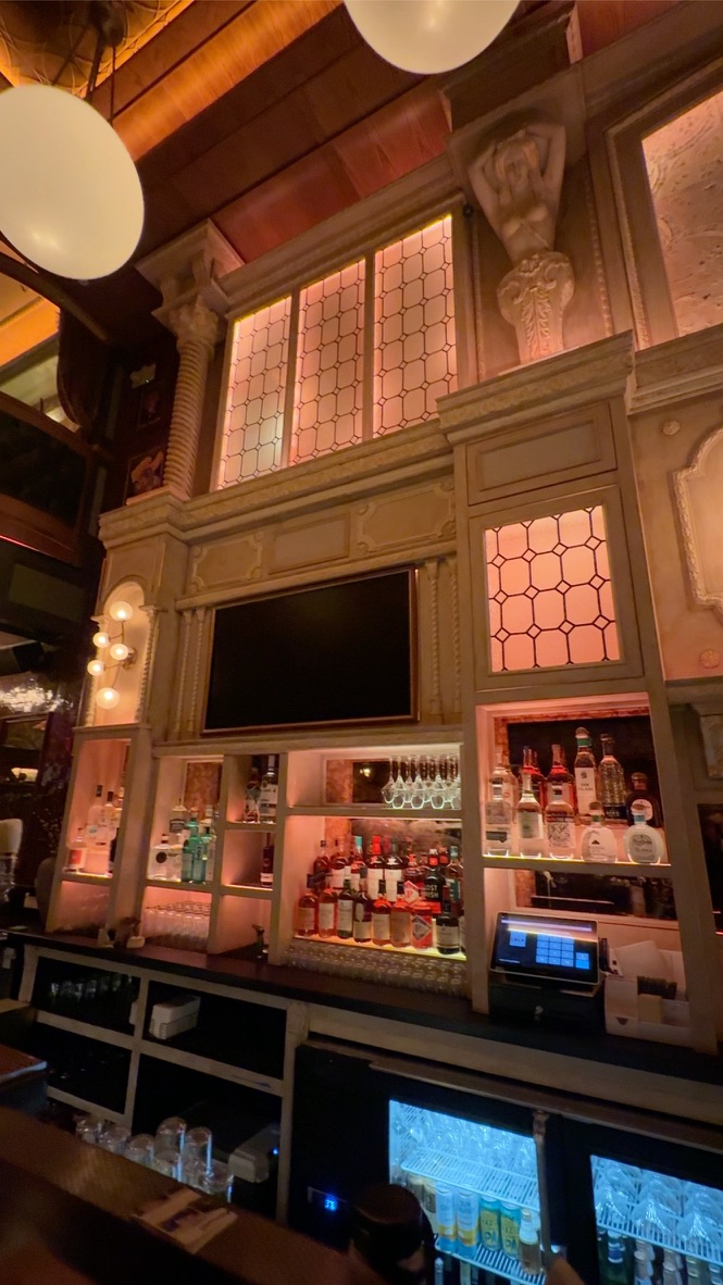 Warm lighting illuminating Bar Lola’s patterned glass, ornate woodwork and bottle shelving